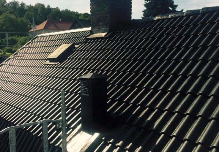 Ett tegeltak med två skorstenar och två takfönster, delvis avgränsat av byggnadsställningar, antyder en pågående takläggning eller takbyte under en klar himmel med träd i bakgrunden.