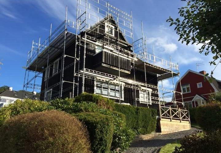 Ett svart hus omgivet av byggnadsställningar, vilket tyder på bygg- eller renoveringsarbete snickeri, med trimmade buskar och en stenlagd gång i förgrunden. Ett svart hus omgivet av byggnadsställningar, vilket tyder på bygg- eller renoveringsarbete snickeri, med trimmade buskar och en stenlagd gång i förgrunden.