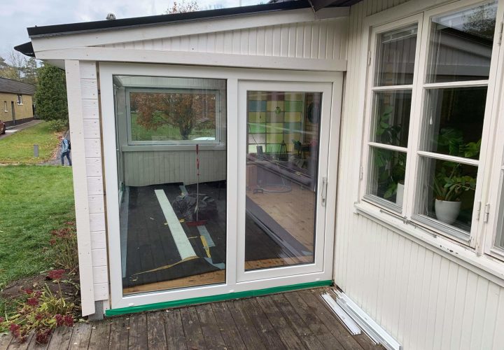 En skjutdörr med vit glasram öppnar sig mot ett trädäck utanför ett hus; inomhus är golvet delvis ofärdigt med synliga byggmaterial, vilket visar på pågående snickeriarbete. En skjutdörr med vit glasram öppnar sig mot ett trädäck utanför ett hus; inomhus är golvet delvis ofärdigt med synliga byggmaterial, vilket visar på pågående snickeriarbete.