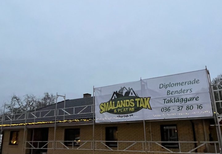 En tegelbyggnad under uppförande med byggnadsställningar och en banderoll för "Smålands Tak & Plåt AB" som annonserar takläggningstjänster.