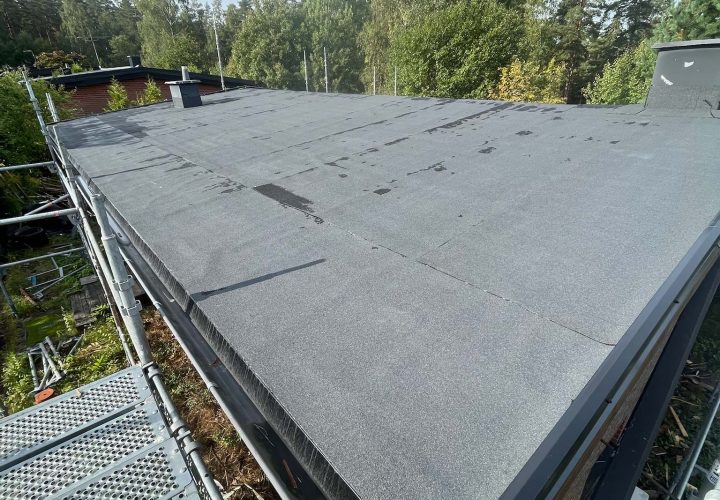 Platt tak täckt med svart takmaterial belyser en pågående takläggning, omgiven av byggnadsställningar och träd i bakgrunden under en klar himmel.